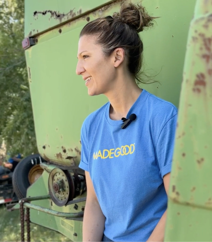 Salma Fotovat in a blue "MADEGOOD" shirt smiles while sitting near a green industrial machine. She has a mic clipped to her shirt, conveying a casual, outdoorsy vibe.